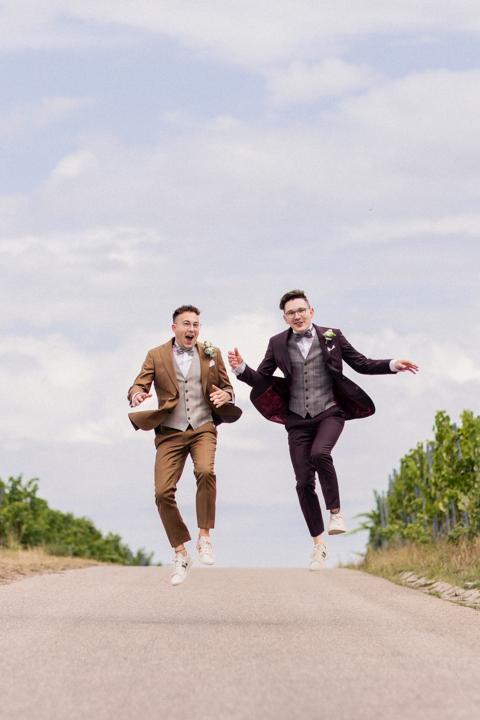 Zwei Bräutigame beim Paarshooting in den Weinbergen, beide springen synchron in die Luft und lachen fröhlich.