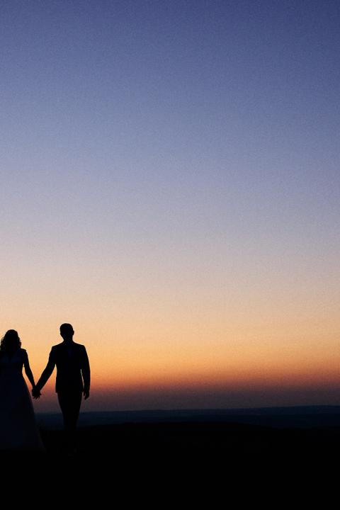 Silhouette eines Brautpaares, das bei Sonnenuntergang Hand in Hand auf einem Hügel spaziert. Der Himmel leuchtet in sanften Farbtönen von Orange und Blau.