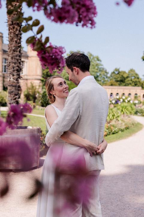 Brautpaar umarmt sich im sommerlichen Schlossgarten zwischen pink blühenden Sträuchern, die Braut schaut lachend zum Bräutigam auf, im Hintergrund Schlossturm und Orangerie