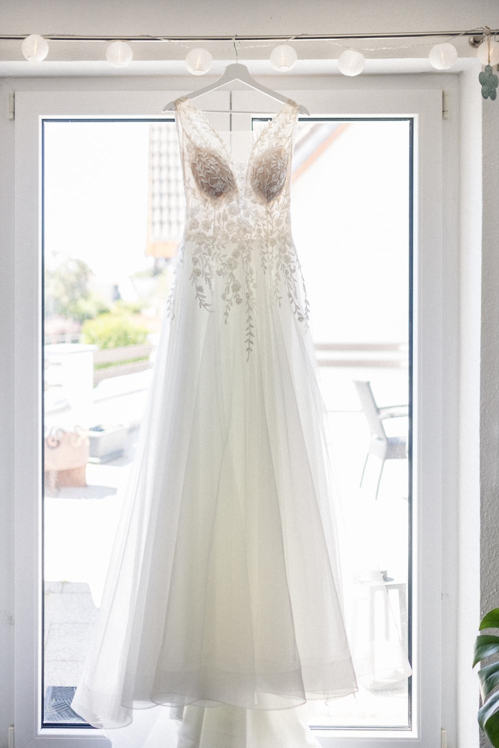 Brautkleid mit Spitzenoberteil und Tüllrock hängt vor dem Fenster am Bügel, Blick auf den Balkon