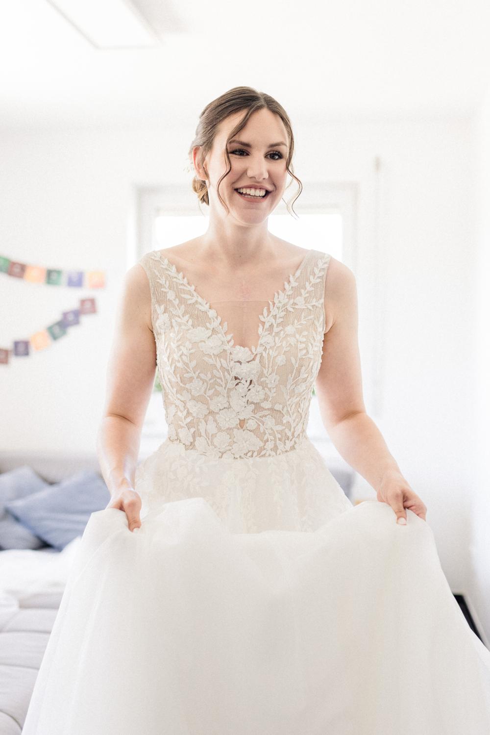 Braut im Spitzenkleid mit V-Ausschnitt hält lachend ihren Tüllrock, helles Wohnzimmer beim Getting Ready