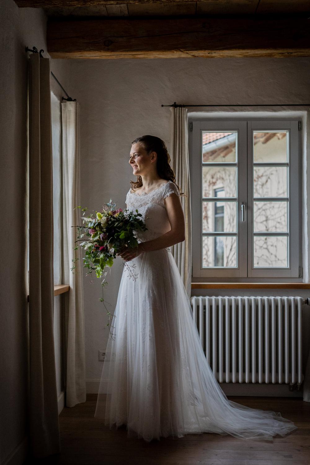 Braut steht mit Brautstrauß im Gegenlicht am Fenster