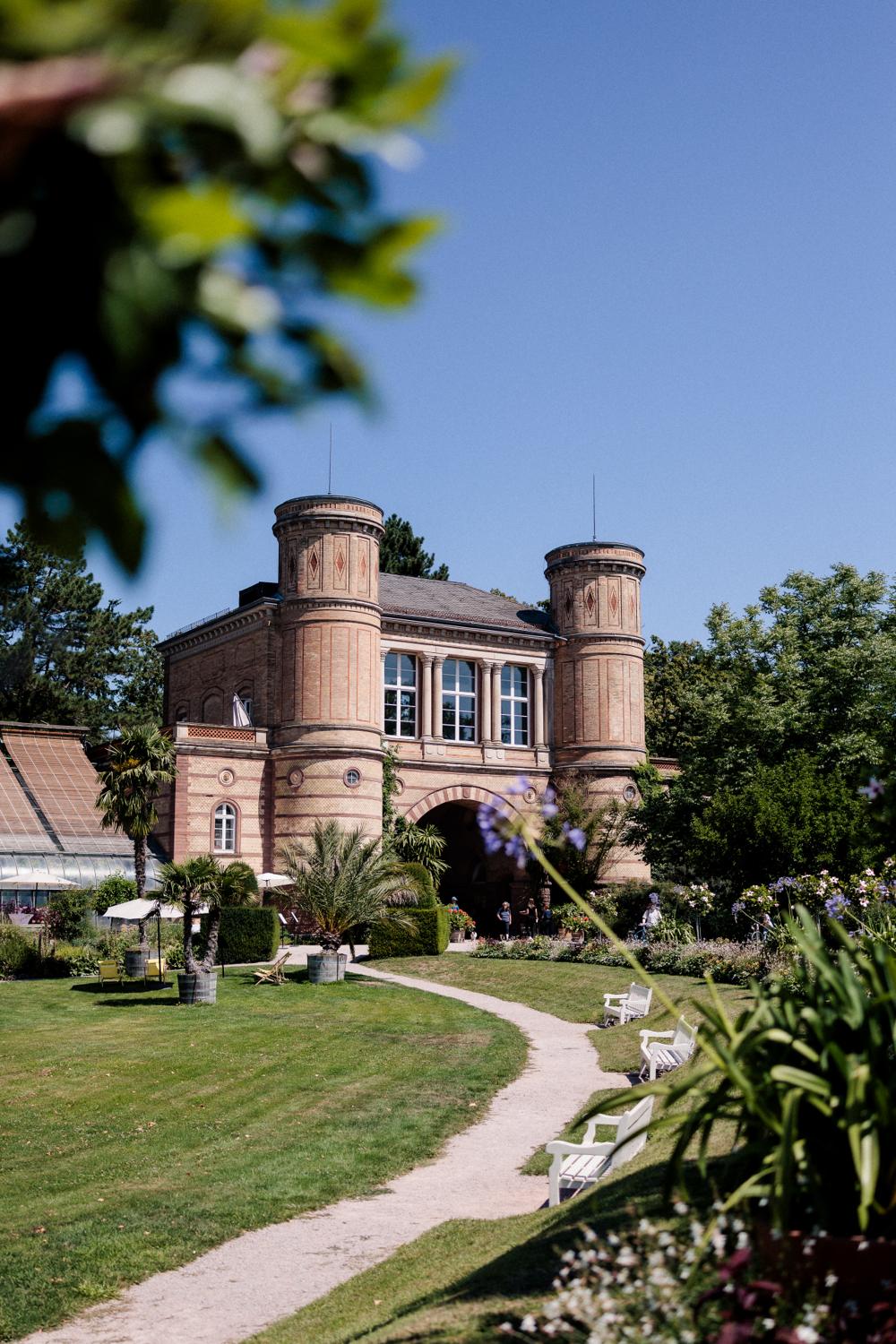 Torbogengebäude des Botanischen Gartens Karlsruhe, gesäumt von Parkbänken und geschwungenem Kiesweg