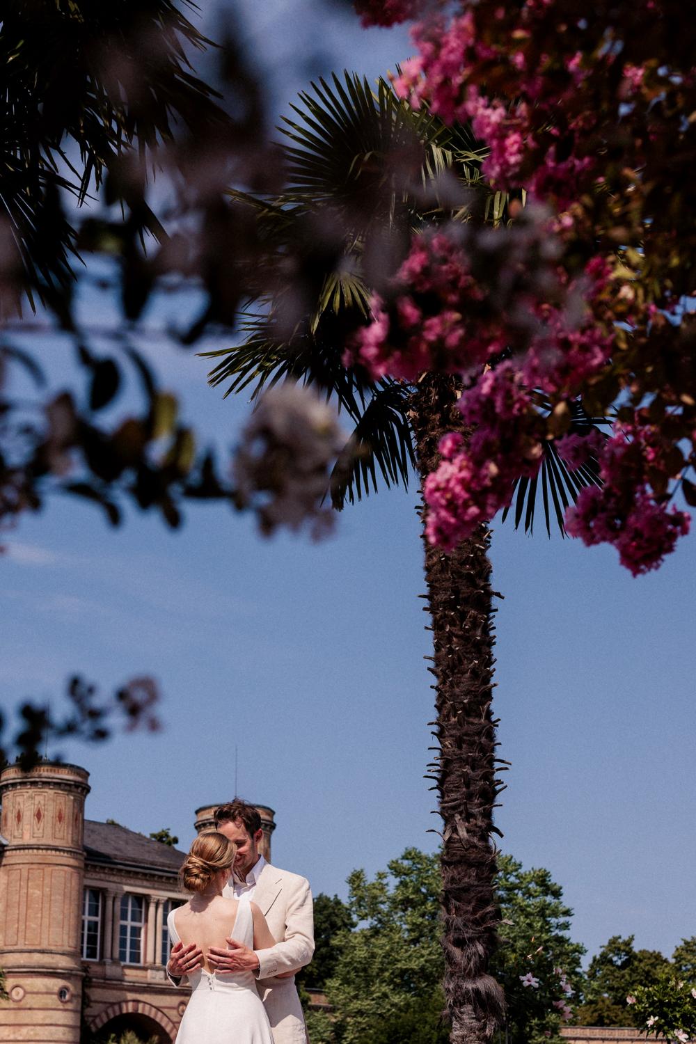 Brautpaar umarmt sich neben einer hohen Palme mit pinken Blüten, ein Turm im Hintergrund