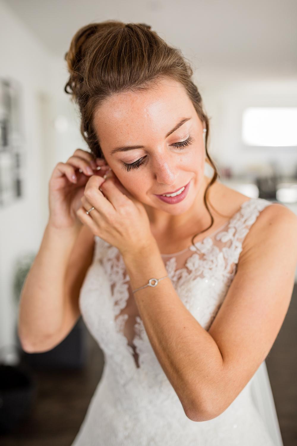 Getting Ready: Braut setzt sich Ohrringe ein, Armband und Spitzenkleid im Vordergrund