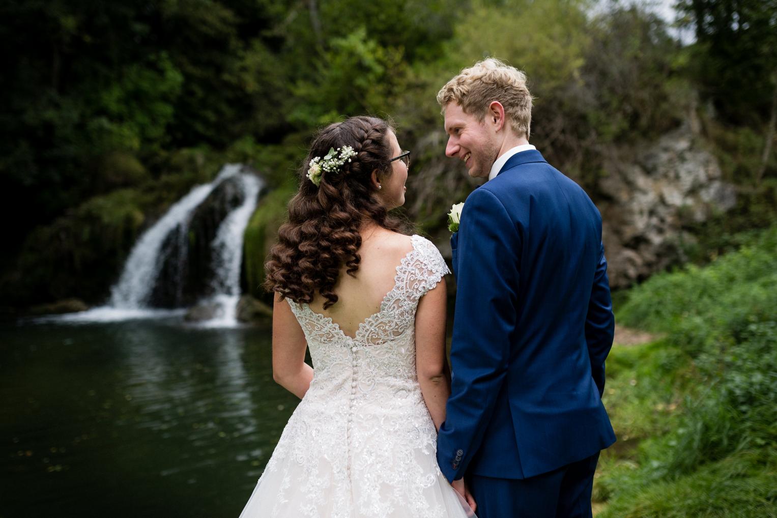Braut von hinten mit Spitzenrücken und Blumenhaarschmuck, Wasserfall im Hintergrund Veringenstadt