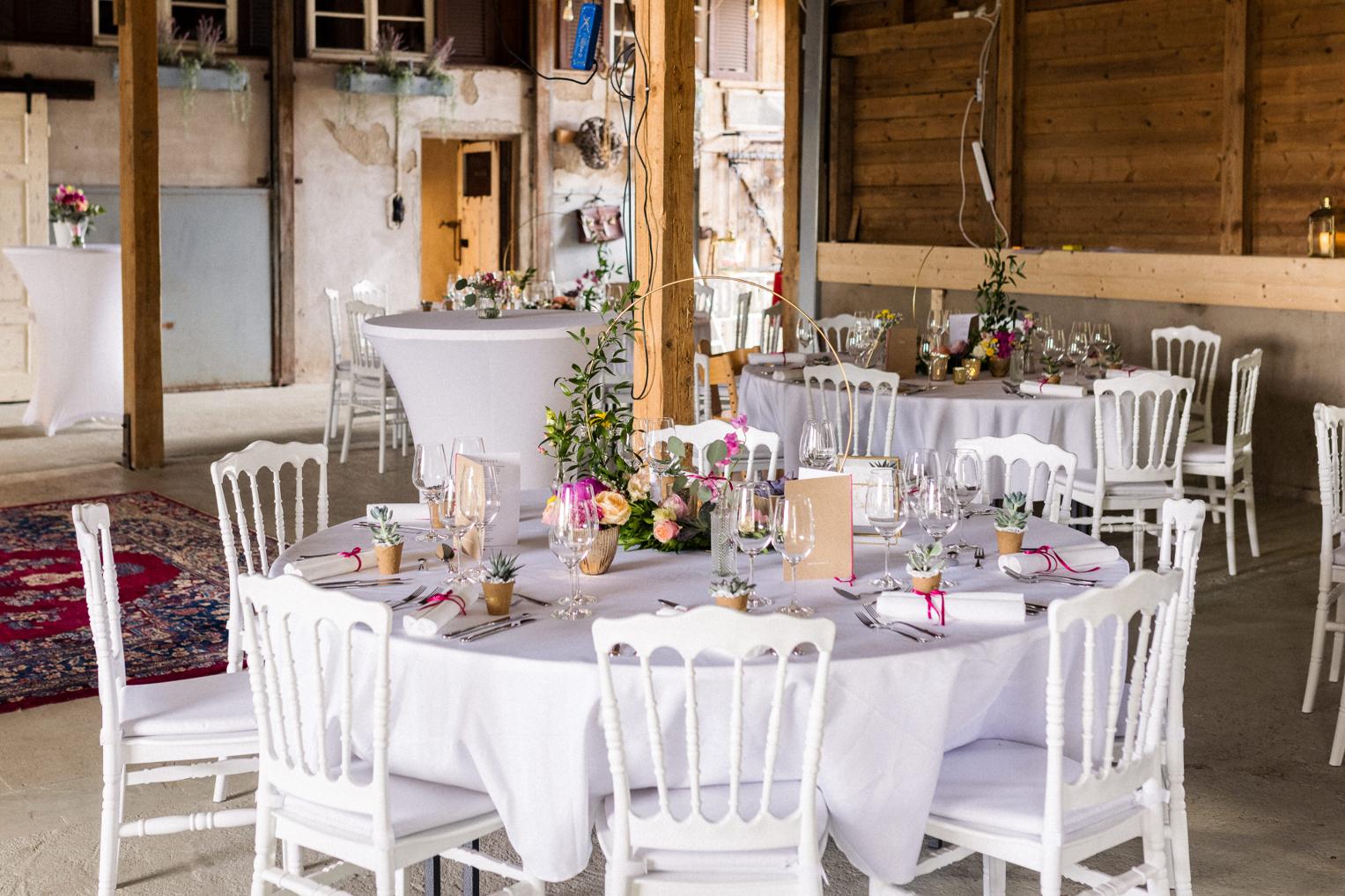 Gedeckte Hochzeitstische mit weißen Stühlen und Blumenarrangements in der Lindenhof-Scheune