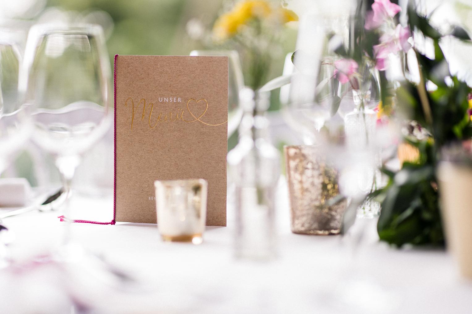 Handgemachte Menükarte mit Goldschrift auf rustikalem Kraftpapier zwischen Hochzeitsblumen