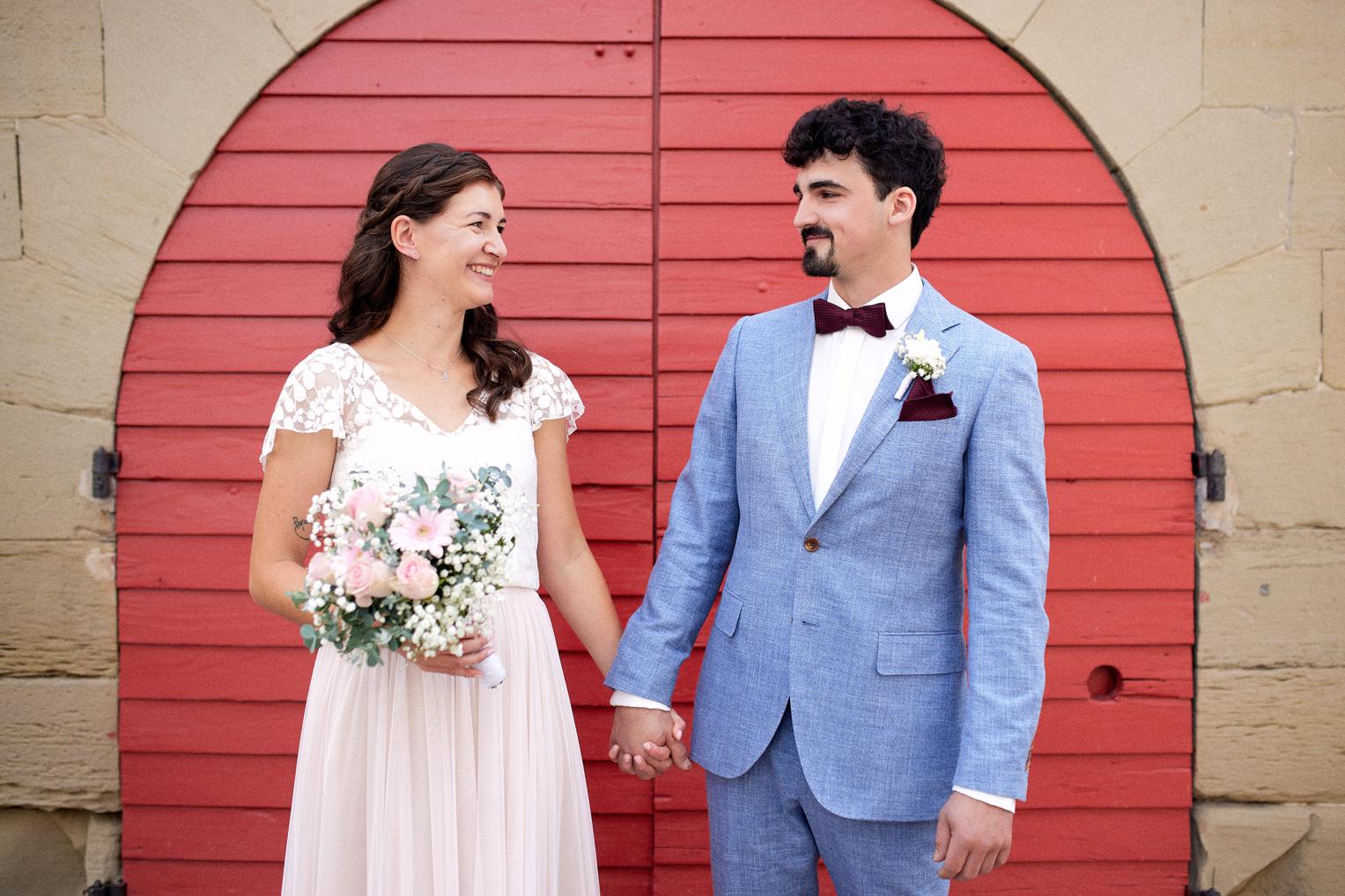 Brautpaar beim Paarshooting vor einer roten Holztür in Herrenberg, Braut mit Blumenstrauß und Bräutigam im eleganten Hochzeitsanzug.