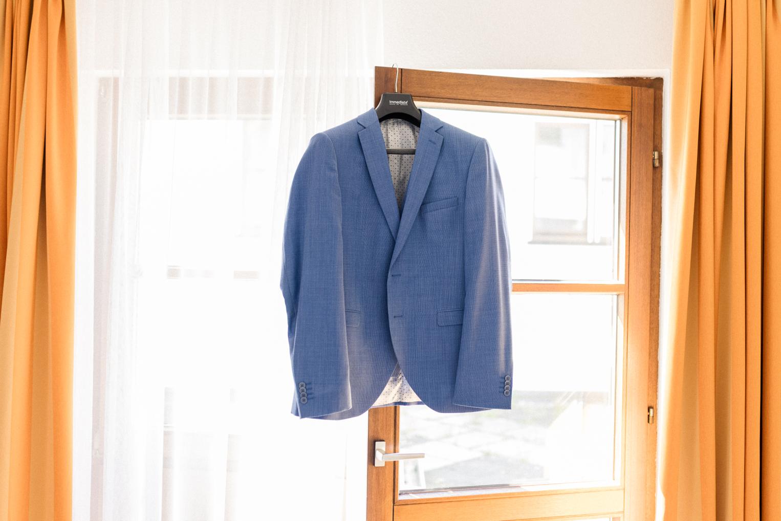 Hellblaues Hochzeitssakko des Bräutigams hängt am Fenster mit orangefarbenen Vorhängen, weiches Tageslicht