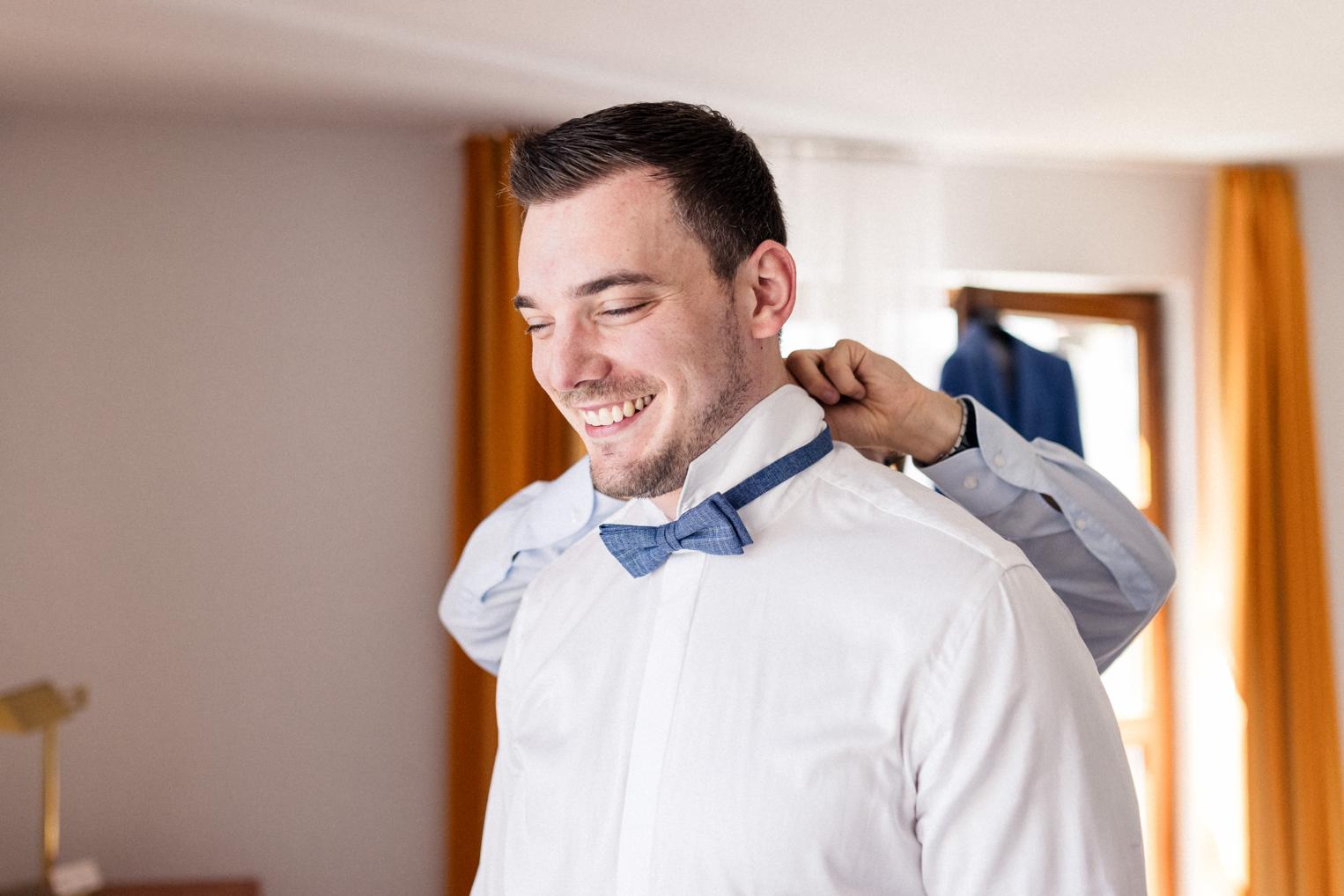 Bräutigam legt lachend seine blaue Fliege an, Getting Ready im hellen Zimmer