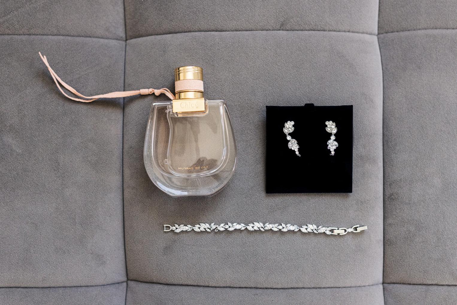 Flatlay der Brautaccessoires – Chloé-Parfum, funkelnde Ohrringe und silbernes Armband auf grauem Samt
