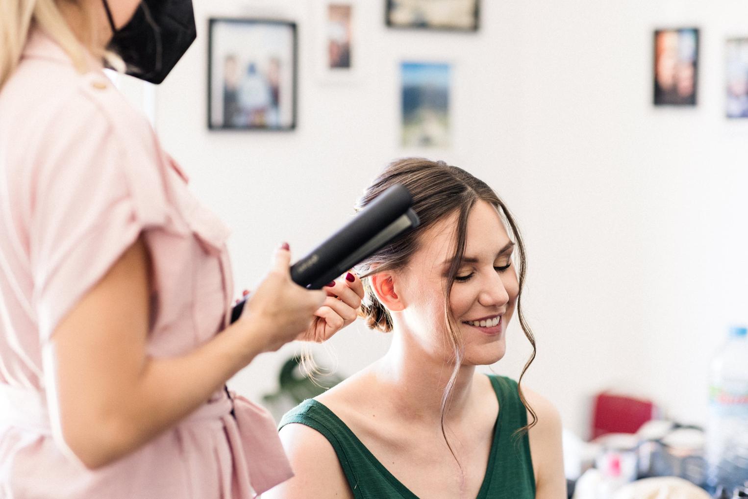 Stylistin legt mit dem Glätteisen die Haare der Braut, Braut lächelt mit geschlossenen Augen