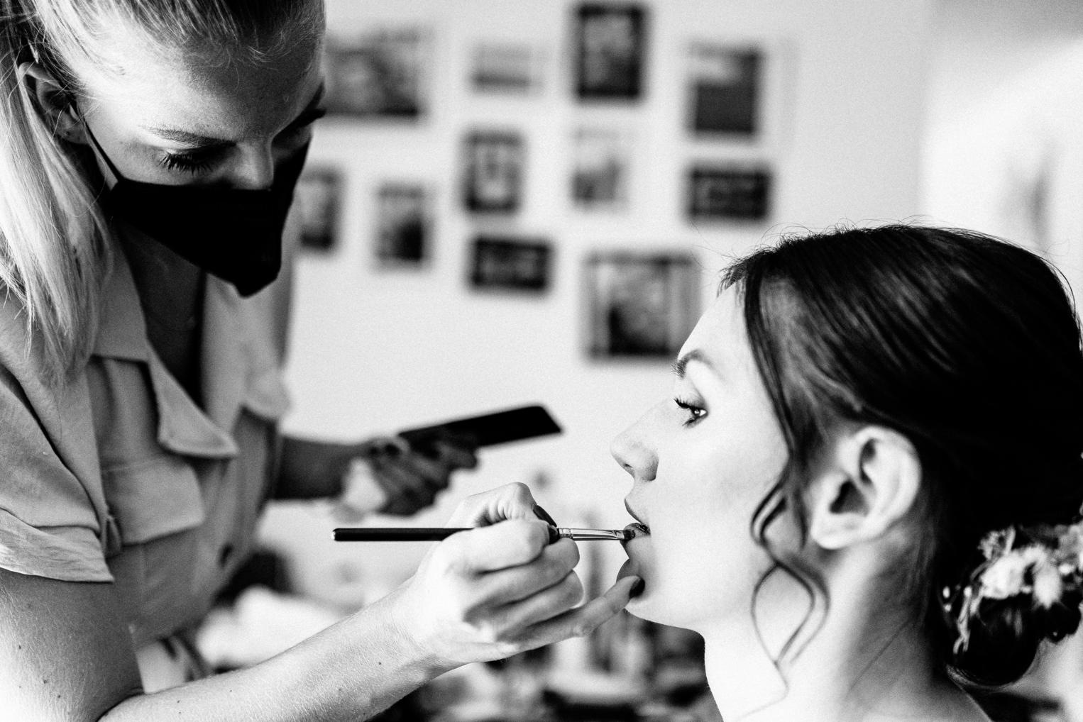 S/W – Make-up-Artistin trägt der Braut Lippenstift auf, Fotowand im Hintergrund