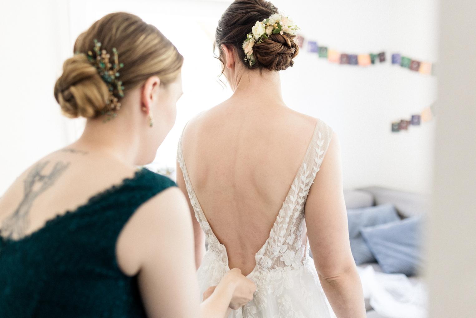 Trauzeugin in grünem Kleid schließt das rückenfreie Spitzenkleid der Braut, beide mit Hochsteckfrisur