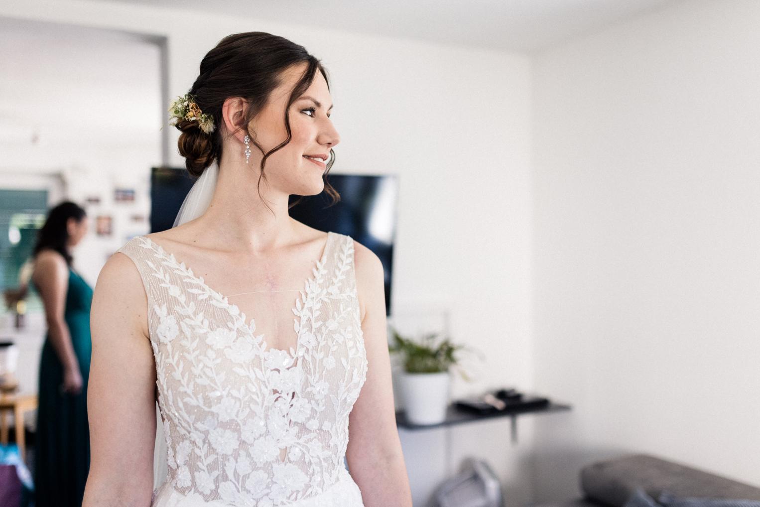 Braut im Spitzenkleid mit Schleier blickt lächelnd zur Seite, Getting Ready im Wohnzimmer