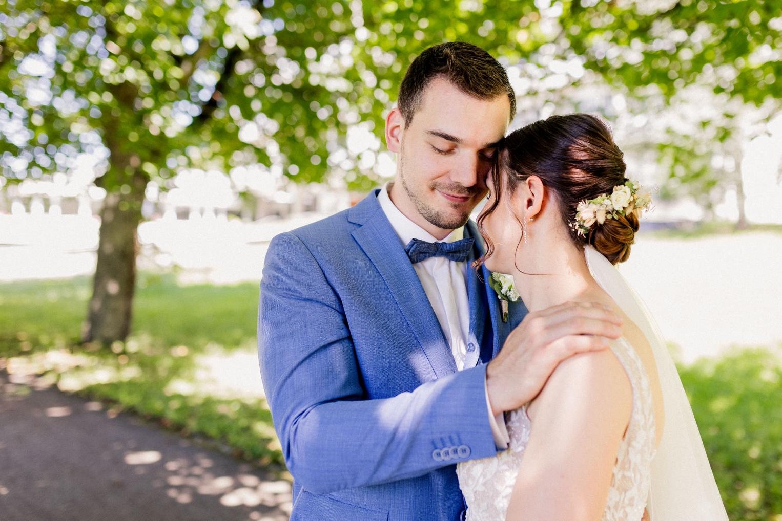 Bräutigam lehnt seine Stirn an die Braut, Hand auf ihrer Schulter, Parkweg im Hintergrund
