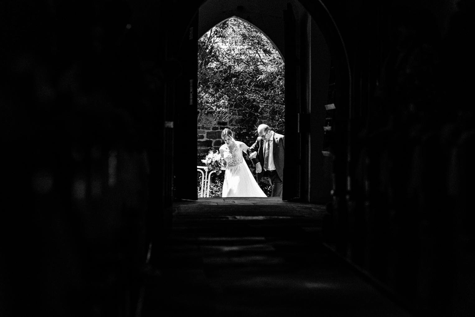 S/W – Braut wird von ihrem Vater durch das Kirchenportal geführt, Blick aus dem dunklen Kirchenraum