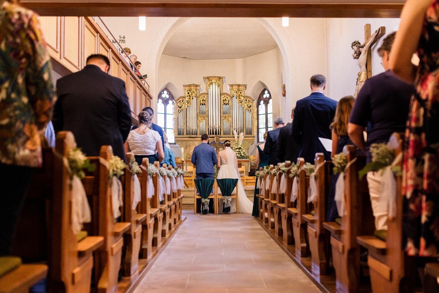 Blick durch das Kirchenschiff zum Altar mit stehenden Gästen, Kirchenbänke mit Blumenschmuck