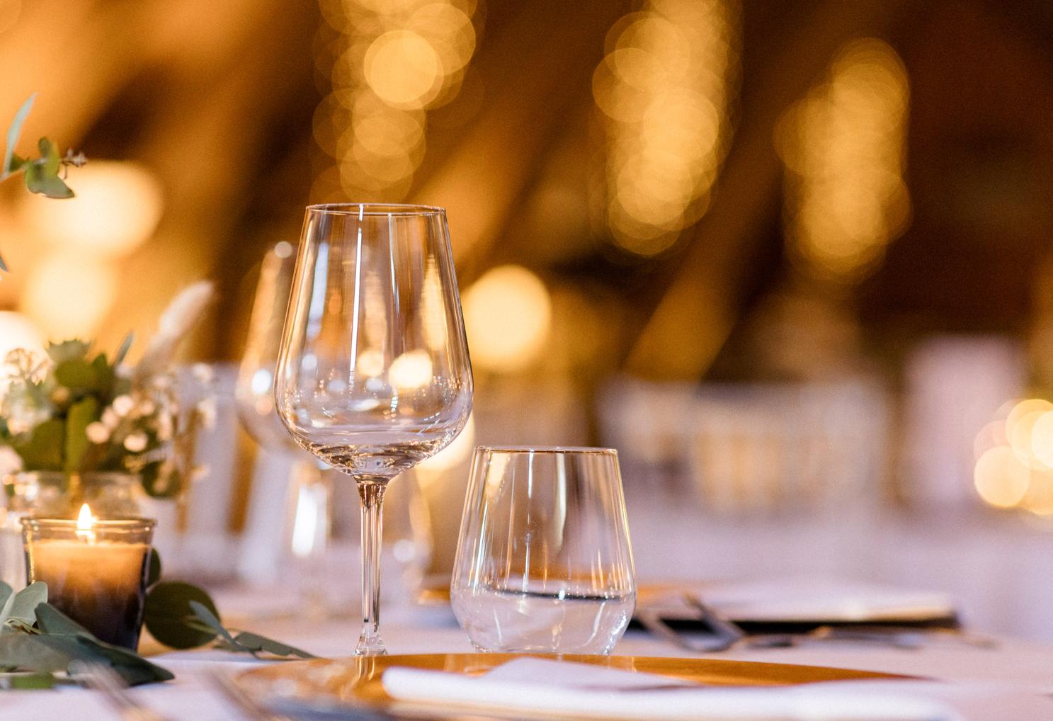 Weinglas und Kerzenlicht auf dem Hochzeitstisch, Lichterketten im Holzgebälk unscharf im Hintergrund