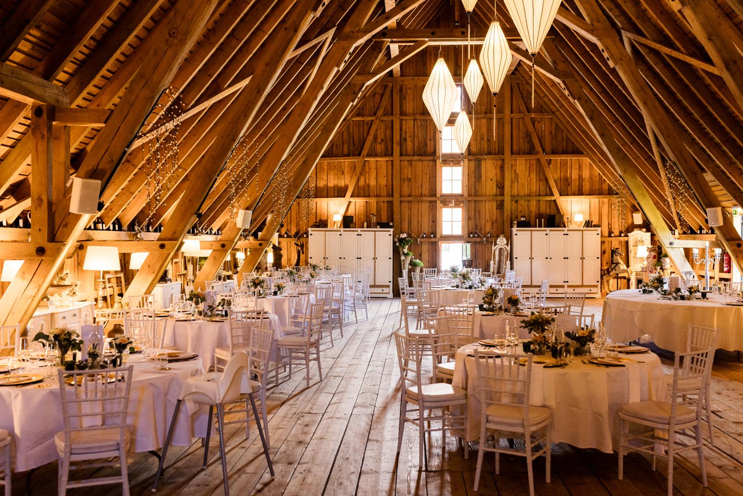 Runde Hochzeitstafeln unter dem Holzgebälk der Scheune im Hofgut Bärenschlössle, Lichterketten und Lampions