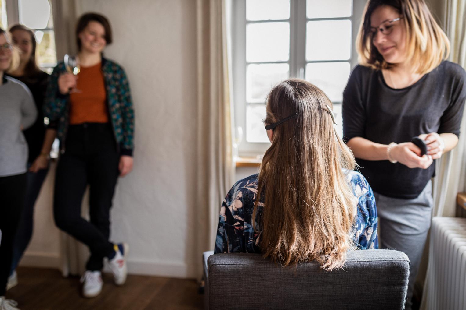 Braut beim Haarstyling während die Trauzeuginnen daneben anstoßen