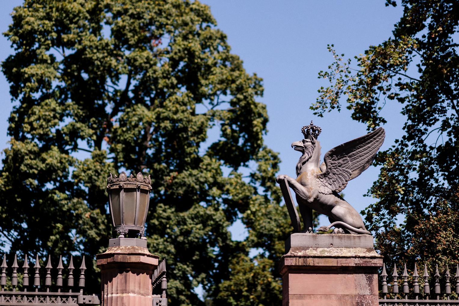 Greif-Skulptur und schmiedeeiserne Laterne am Toreingang des Botanischen Gartens Karlsruhe vor dichten Bäumen