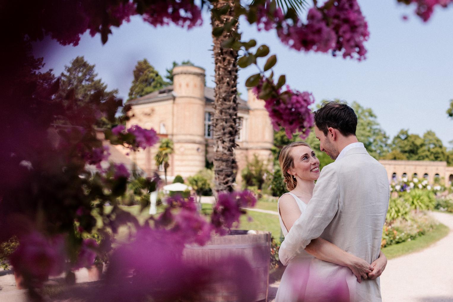 Umarmung des Paares vor dem Torbogengebäude, eingerahmt von pinken Blüten – Botanischer Garten Karlsruhe