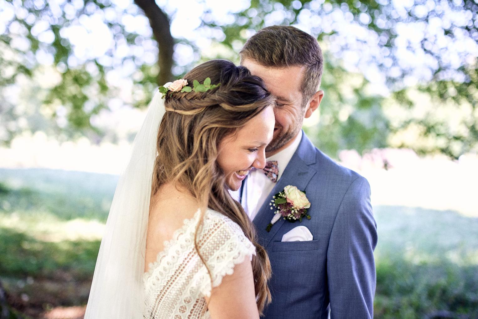 Braut mit Blumenkranz lacht an Bräutigams Schulter beim First Look