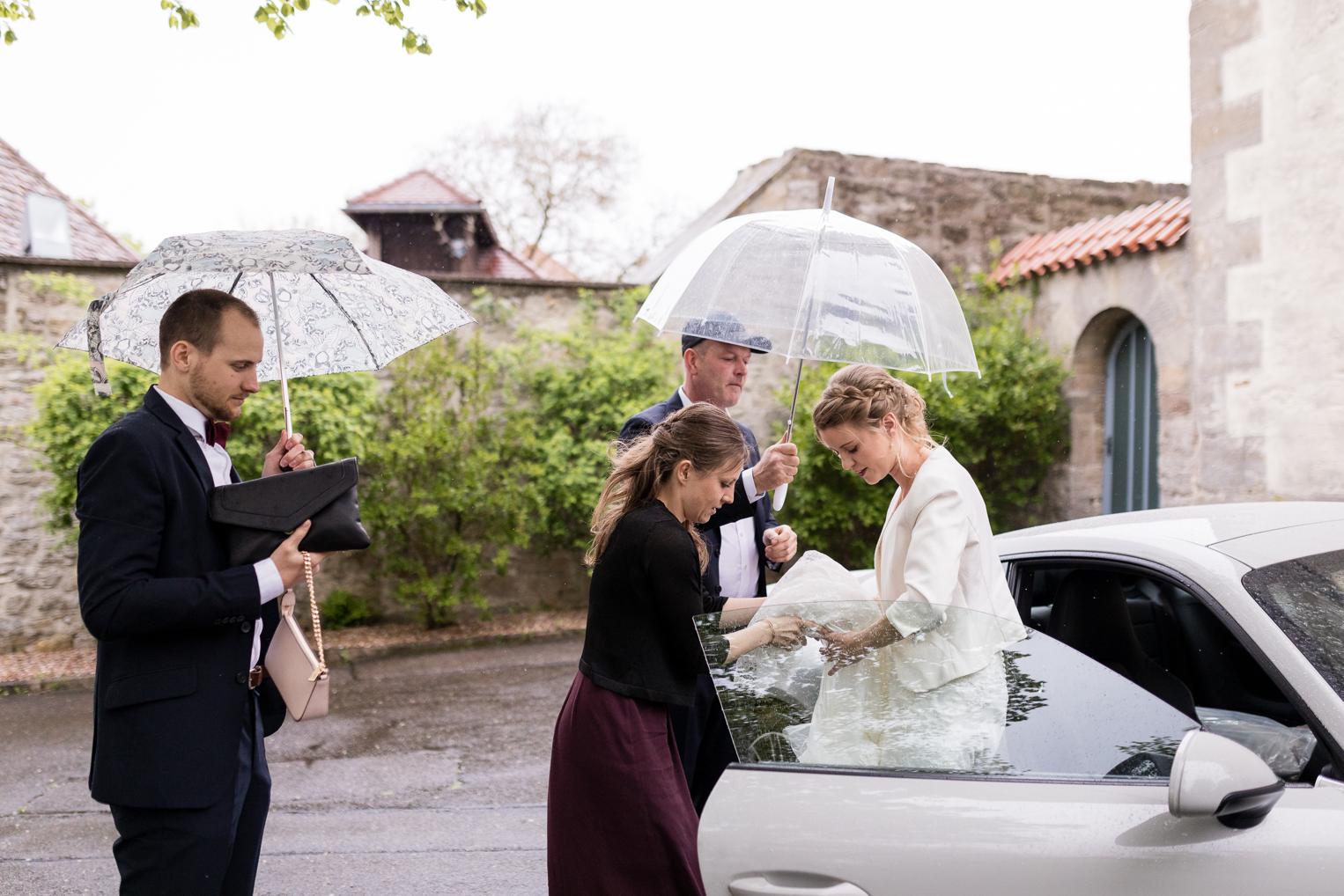 Braut steigt unter Regenschirmen aus dem Auto vor der Kirche in Gärtringen