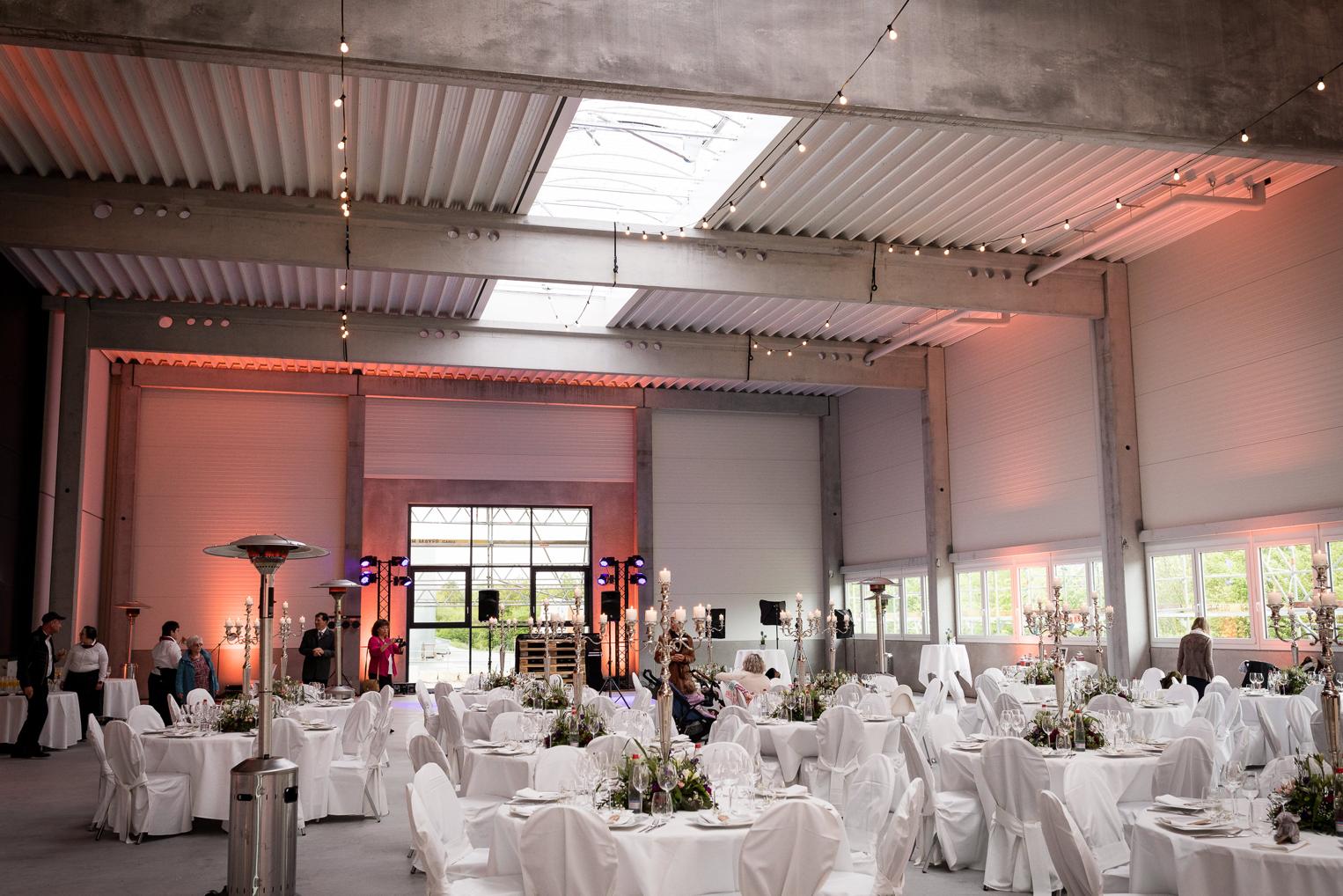 Festlich dekorierter Hochzeitssaal mit weißen Tischrunden, Lichterketten und Industriecharme