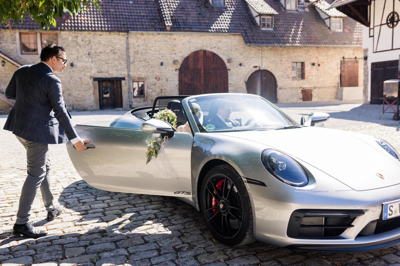 Trauzeuge öffnet der Braut die Tür des Porsche am Landgut