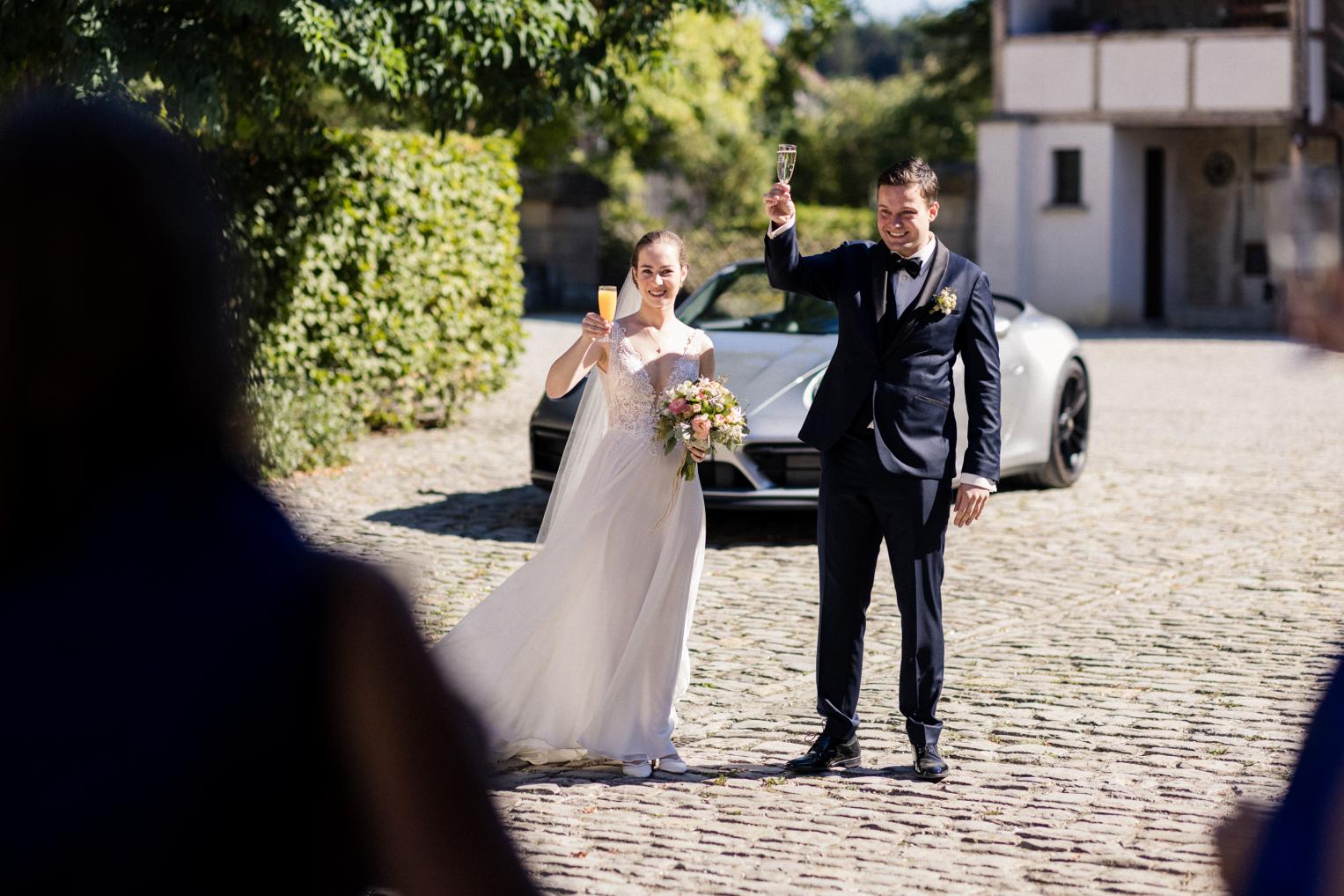 Brautpaar stößt mit Sekt vor dem Hochzeitsauto am Landgut an