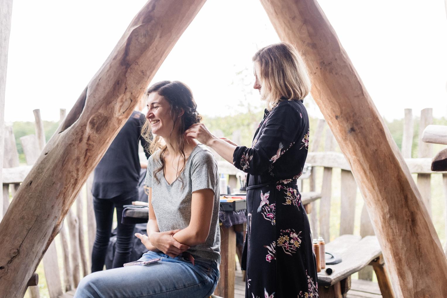Friseurin steckt der Braut lächelnd die Haare auf der Holzterrasse des Baumhaushotels