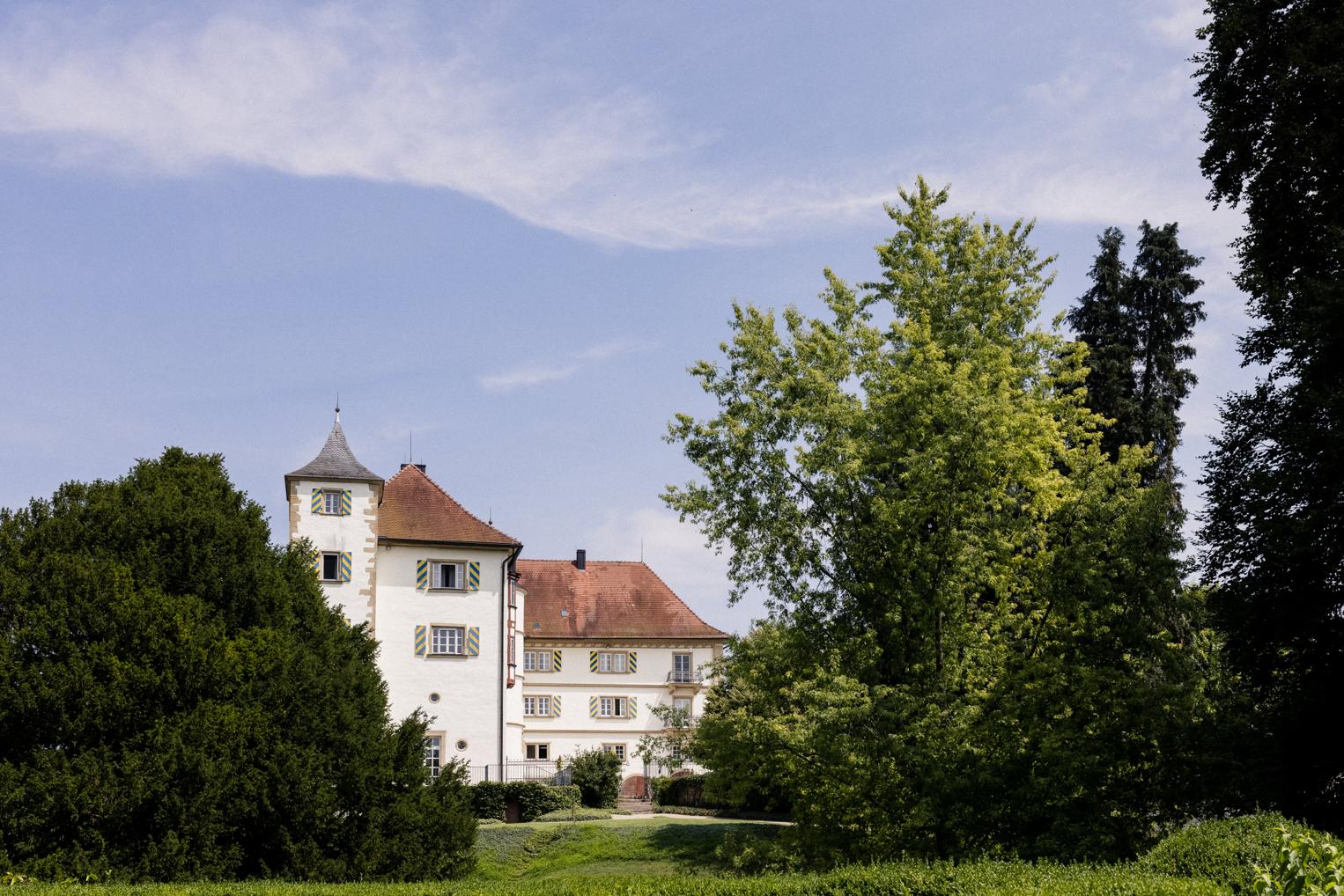 Schloss Neuhaus bei Sinsheim hinter alten Bäumen – weiße Fassade mit Turmhaube unter sommerlichem Himmel