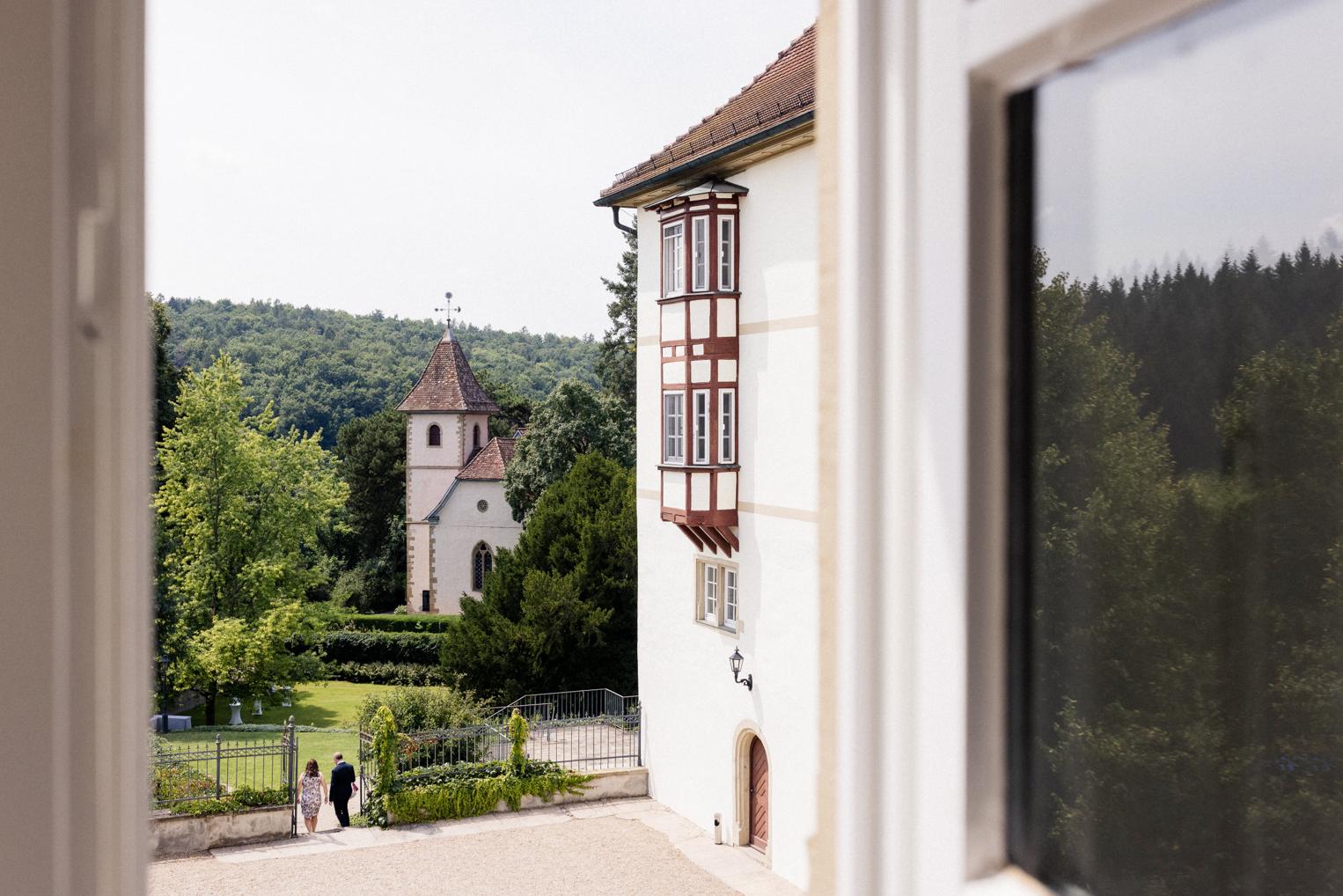 Blick aus einem Schlossfenster auf die Schlosskirche und den Fachwerkerker, im Park darunter zwei Hochzeitsgäste
