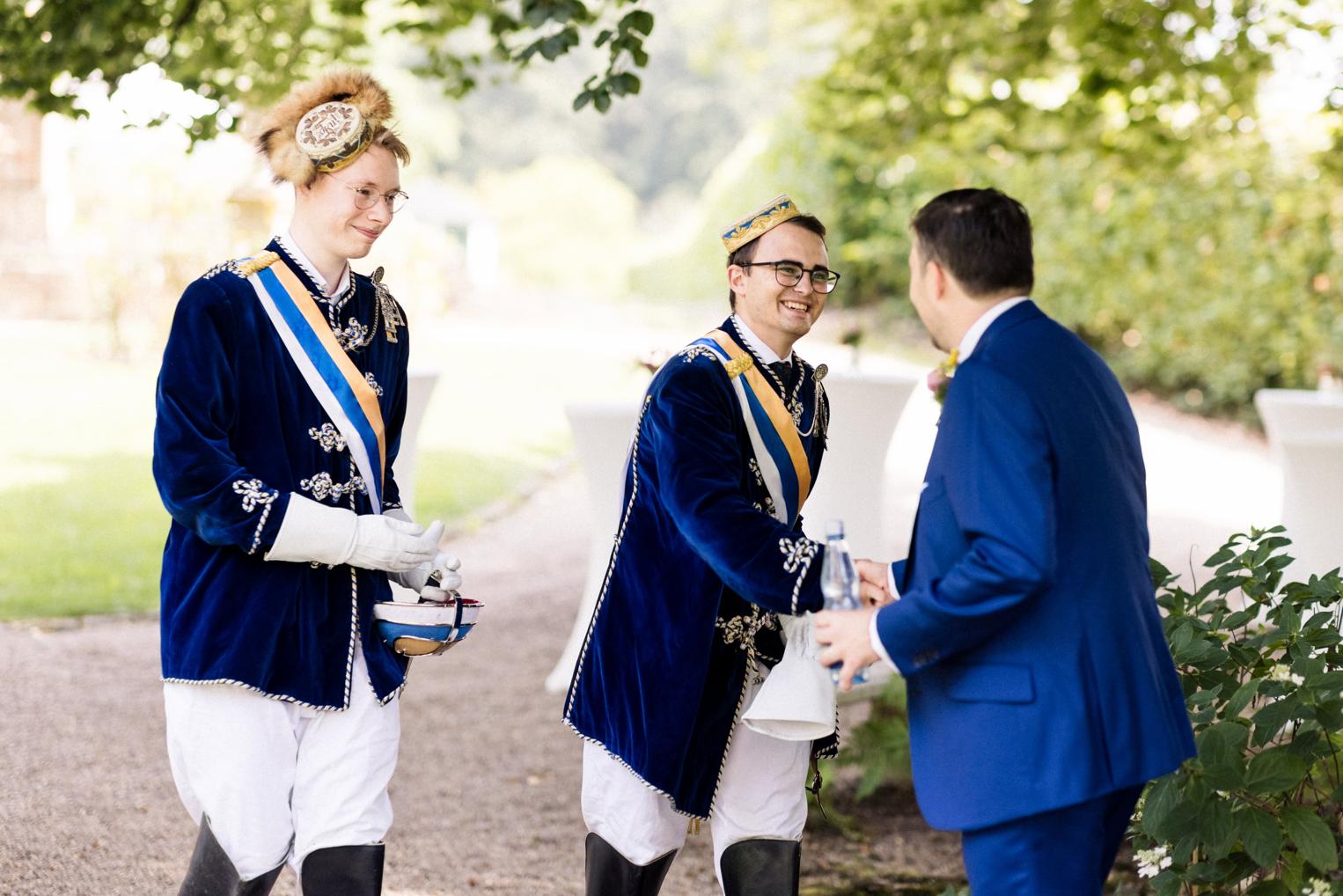 Zwei Chargierte in blauen Couleurröcken und Stulpenstiefeln im Gespräch mit einem Hochzeitsgast im Schlosspark