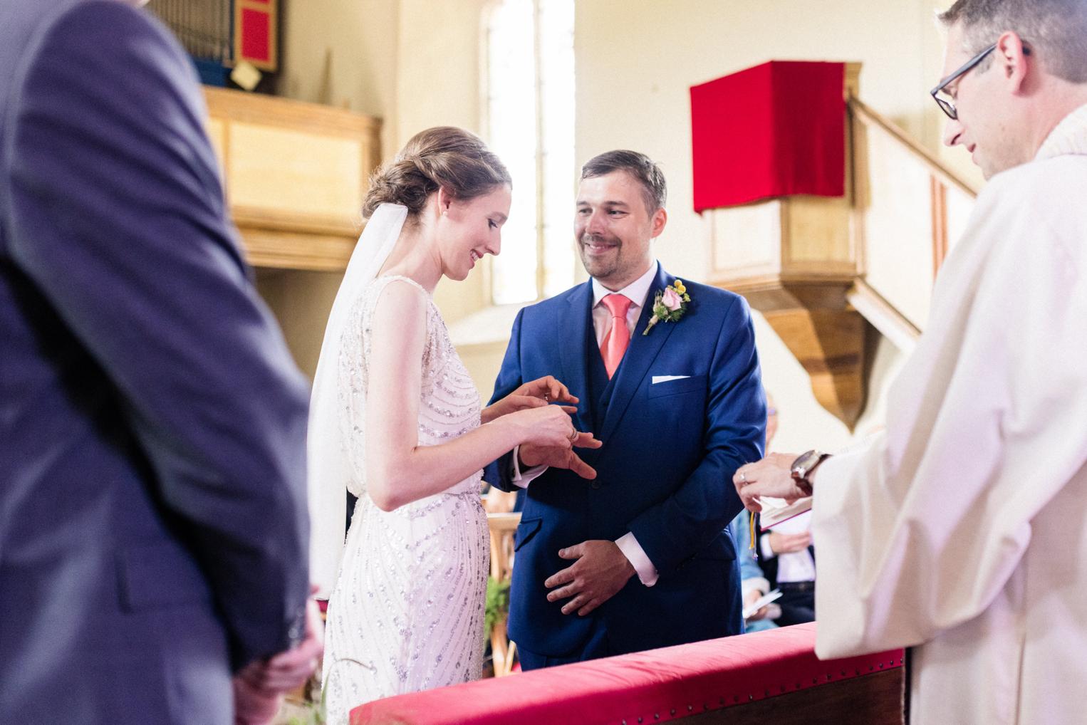 Ringtausch am Altar: Die Braut streift dem Bräutigam den Ehering an, der Pfarrer schaut zu