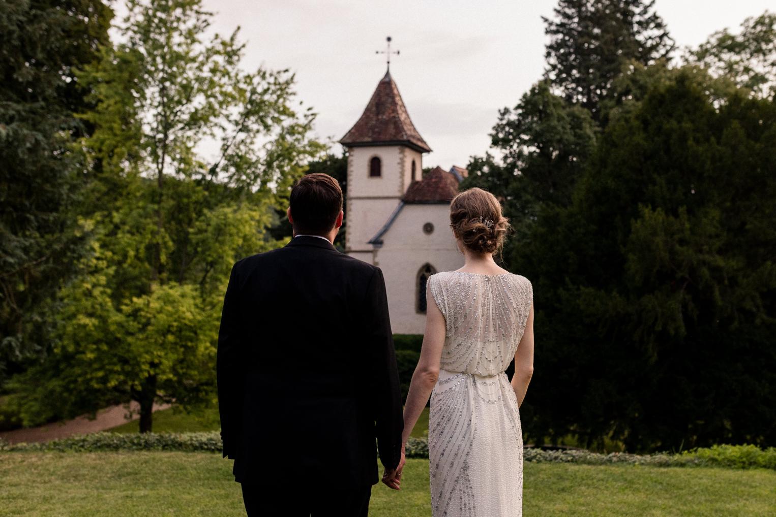 Brautpaar von hinten, Hand in Hand, mit Blick auf die kleine Schlosskirche im Park
