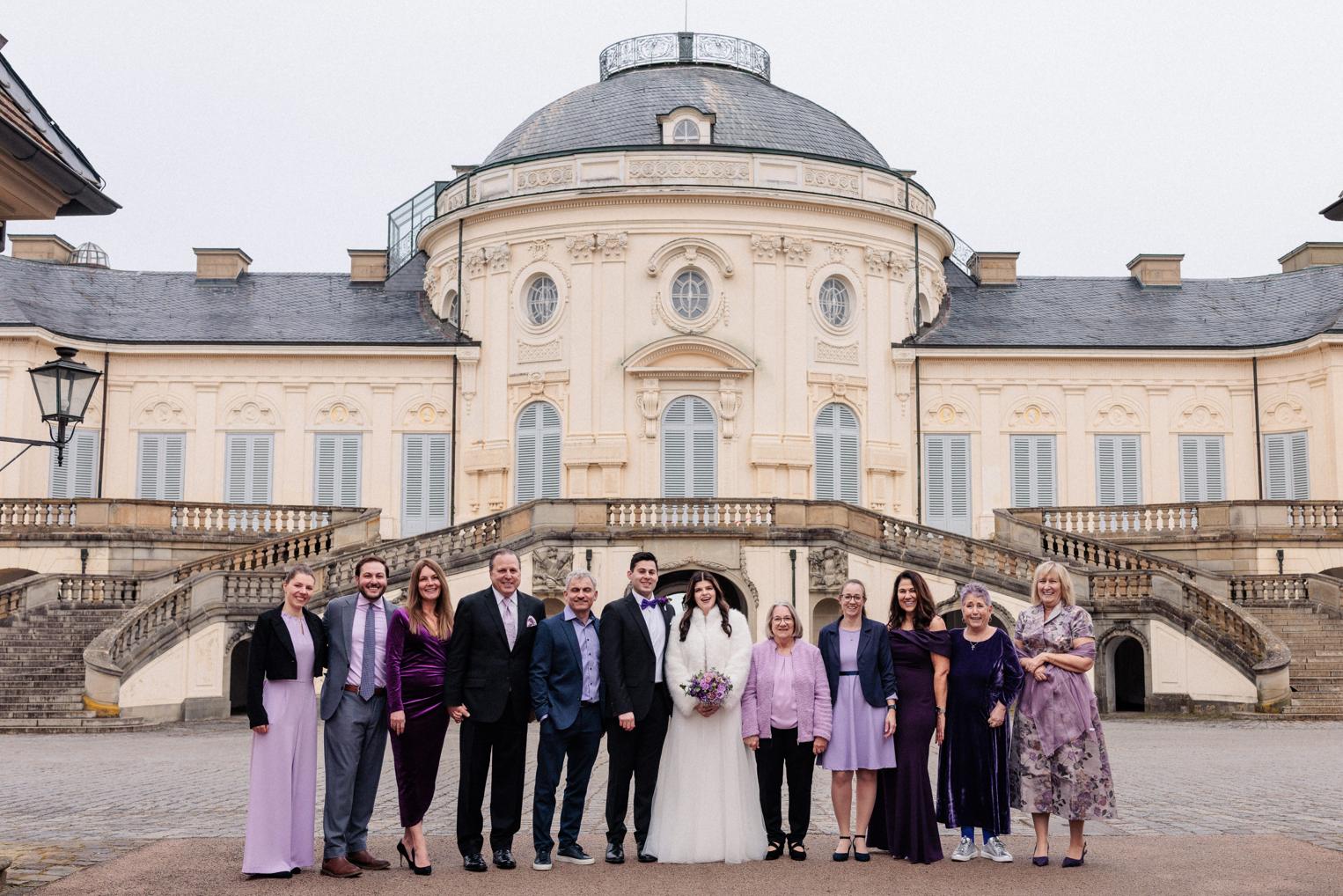 Großes Familien-Gruppenfoto vor der Freitreppe von Schloss Solitude, Brautpaar in der Mitte umgeben von Angehörigen in Lila-Tönen