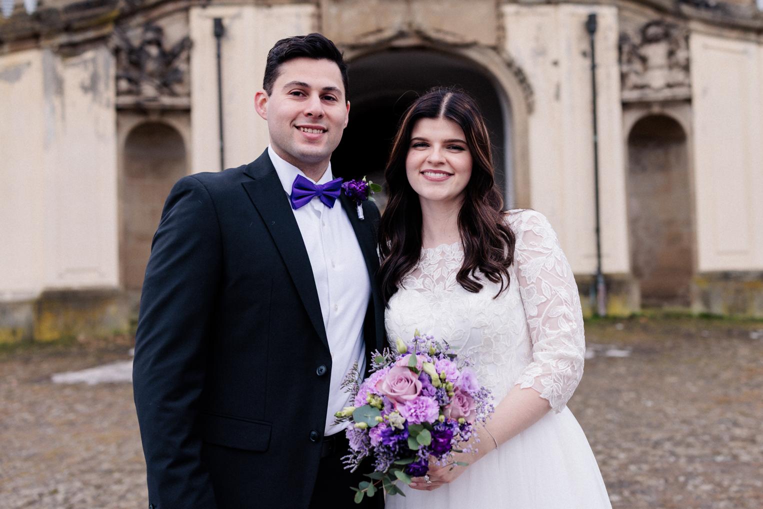 Lächelndes Brautpaar vor dem Torbogen von Schloss Solitude, Braut im Spitzenkleid mit Brautstrauß aus lila Rosen