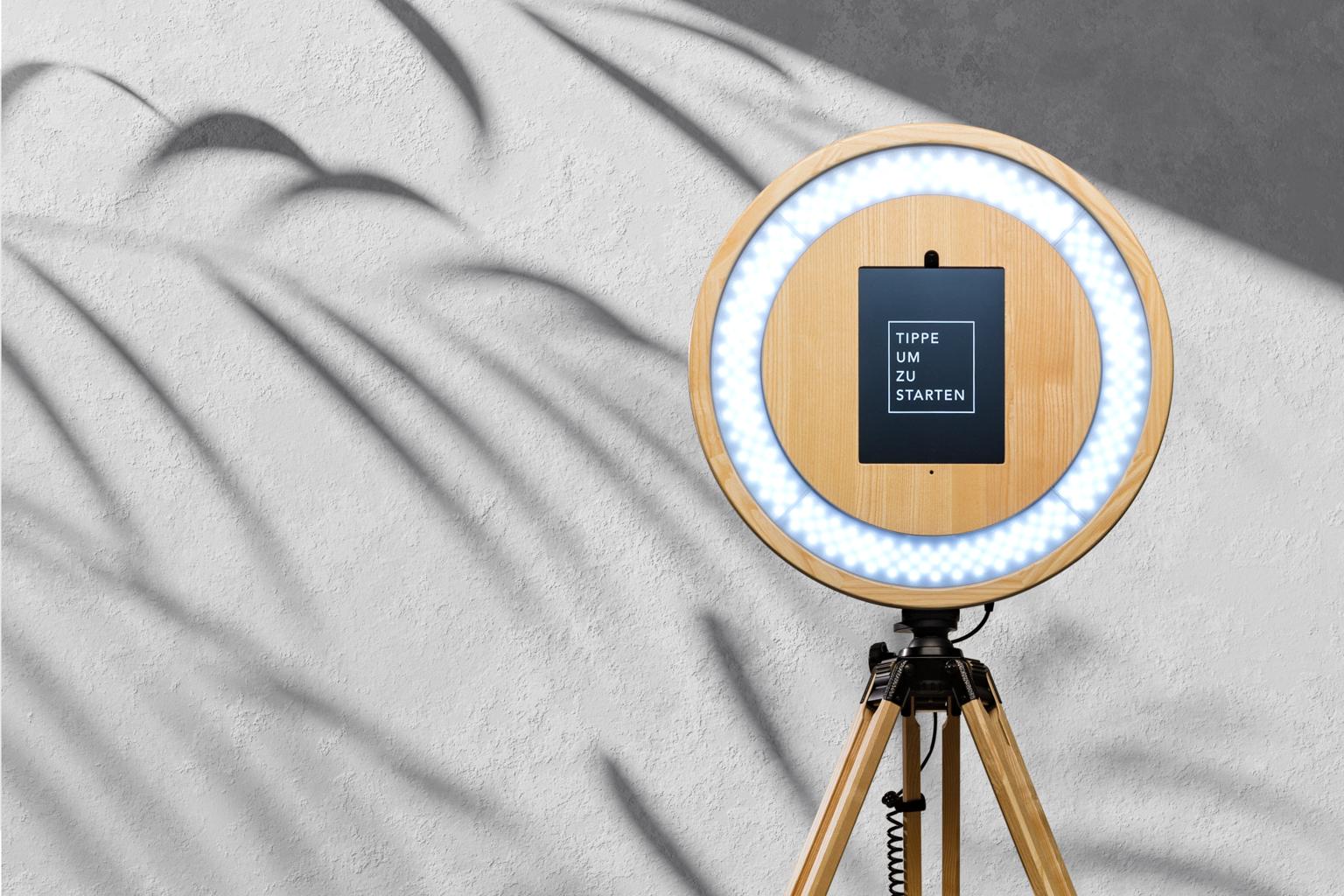 Eine edle Holz-Fotobox für Eure Hochzeit: Handgefertigtes, helles Holz in sanfter Maserung, umrahmt den integrierten Touchscreen; das ringförmige Licht schafft eine warme, natürliche Atmosphäre und steht auf einem stabilen Dreibein.