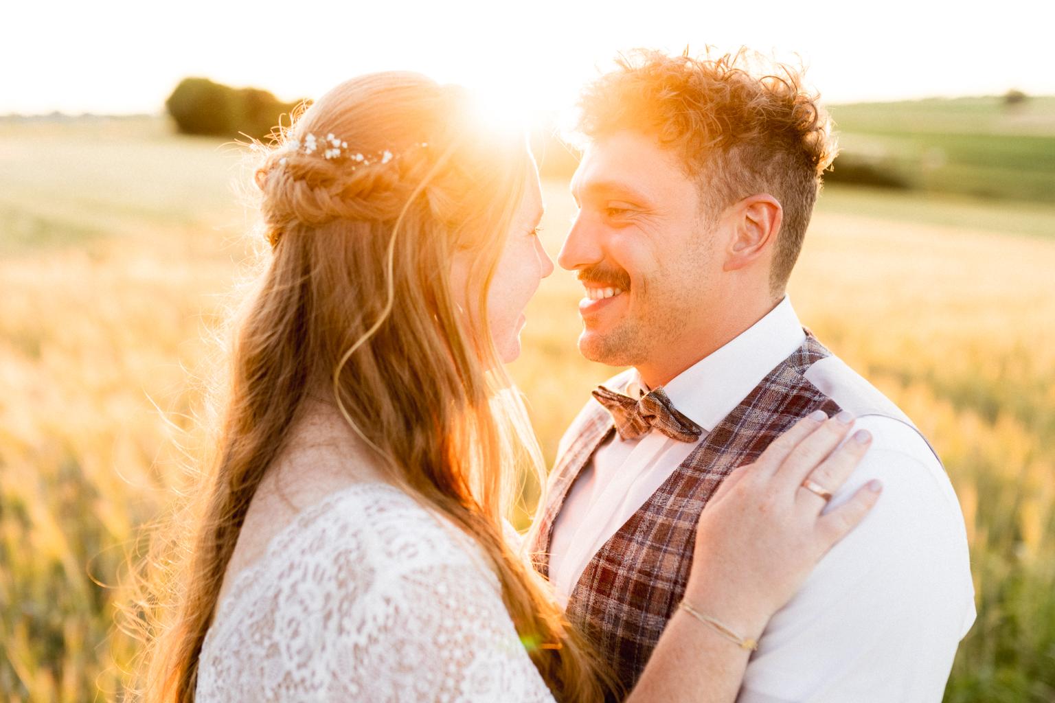 Brautpaarshooting beim Sonnenuntergang: Braut im Spitzenkleid mit locker geflochtenem Haar und Bräutigam in kariertem Gilet und Fliege stehen in einem goldenen Weizenfeld bei untergehender Sonne und lächeln sich verliebt an.