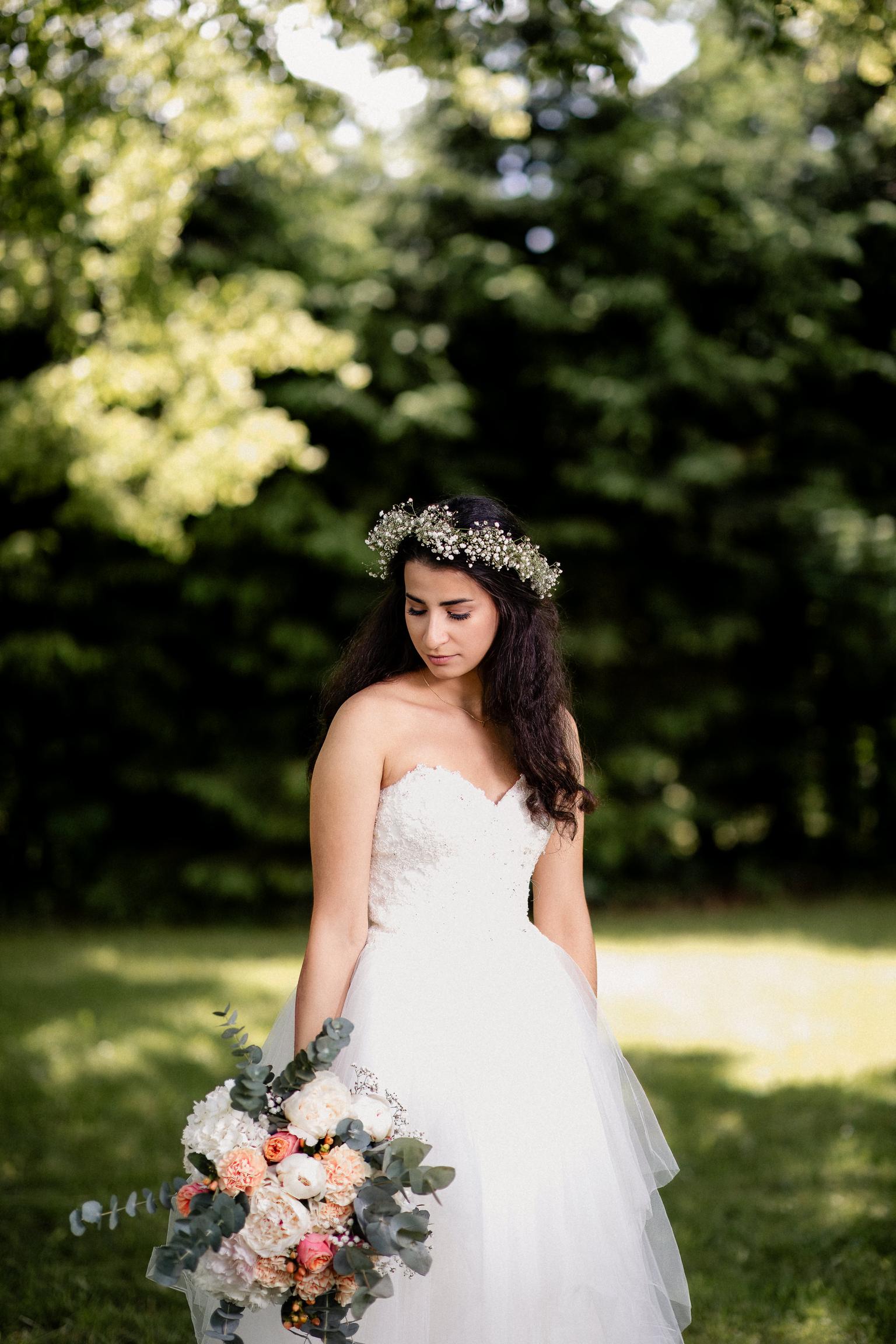 Braut in einem weißen Hochzeitskleid mit Blumenkranz im Haar, posiert mit einem eleganten Brautstrauß in einer sonnigen Gartenkulisse.