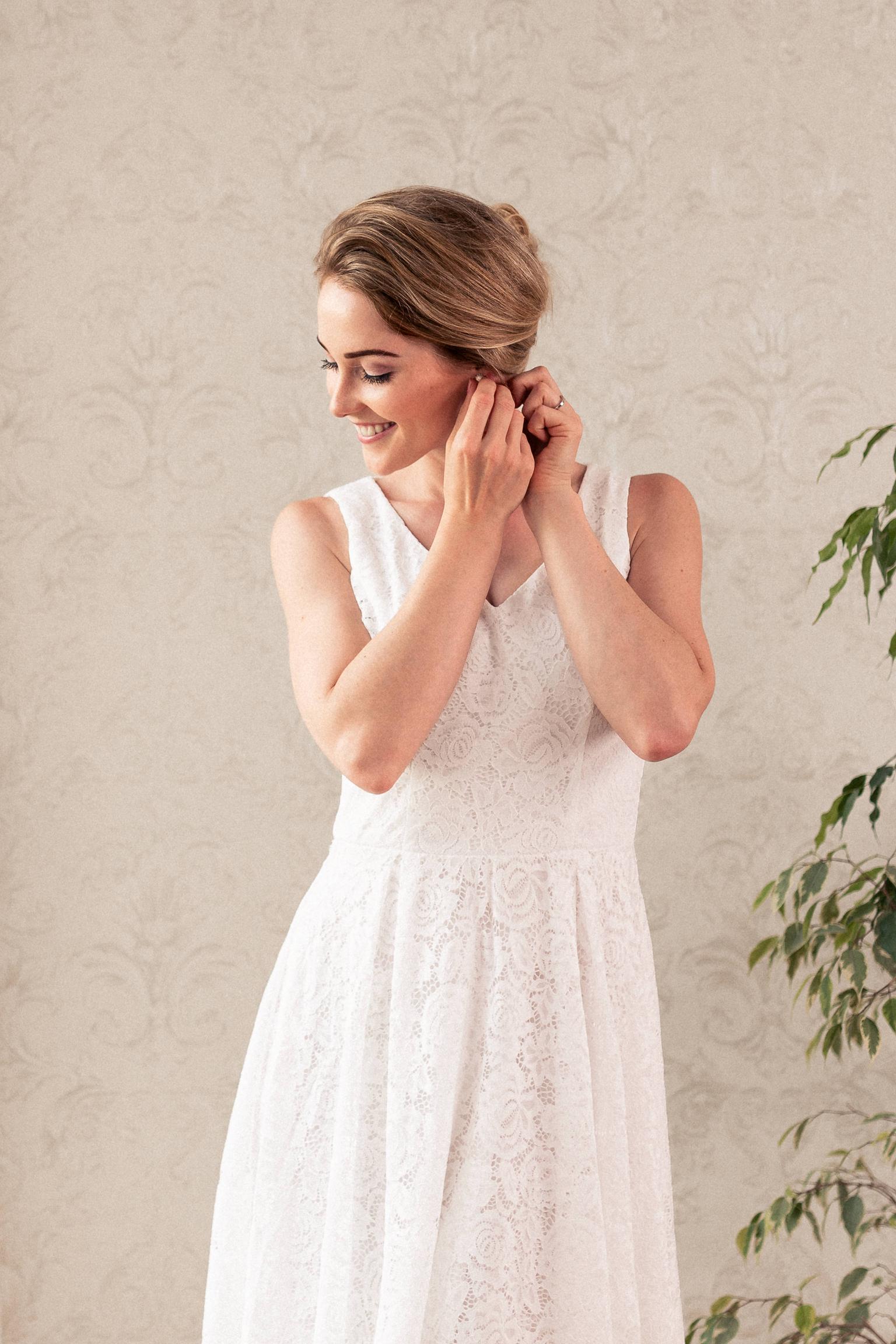 Braut in elegantem Spitzenkleid, die lächelnd ihre Ohrringe anlegt, vor einer dezenten Tapete mit Pflanzen im Hintergrund.