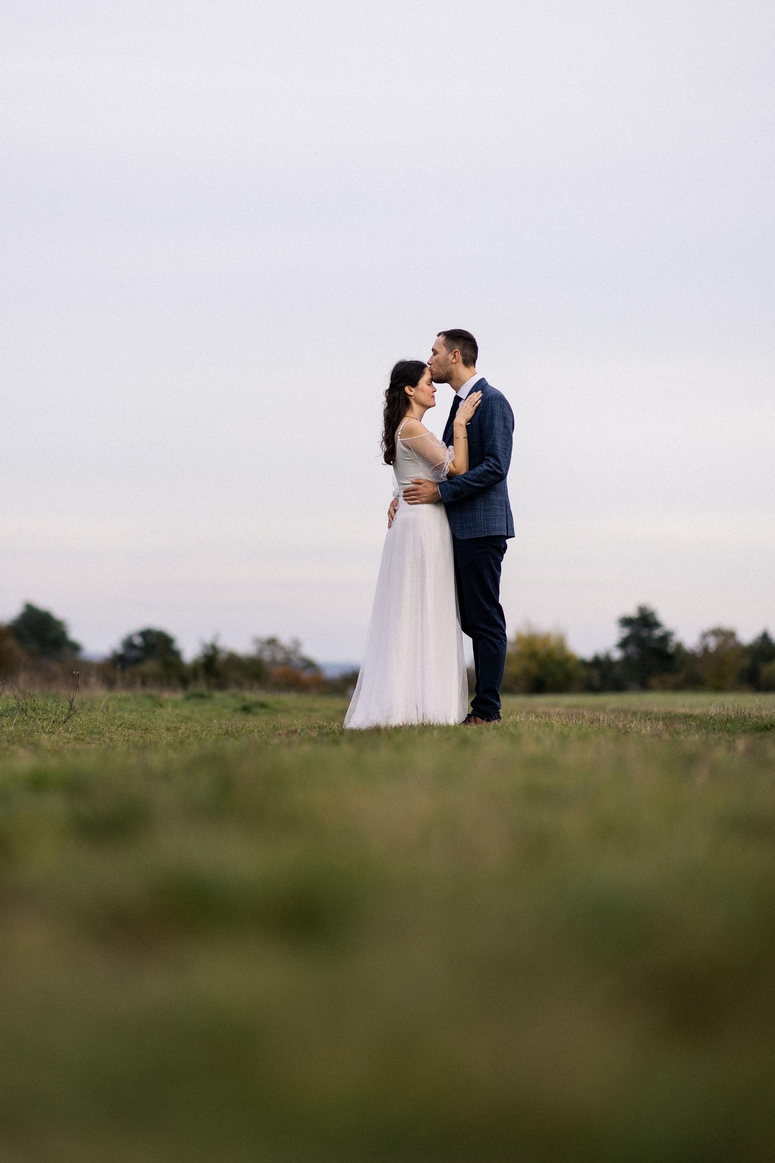 Braut und Bräutigam stehen inmitten einer weiten Wiese im sanften Abendlicht und tauschen einen zarten Kuss, sie im fließenden Kleid, er im eleganten Anzug.