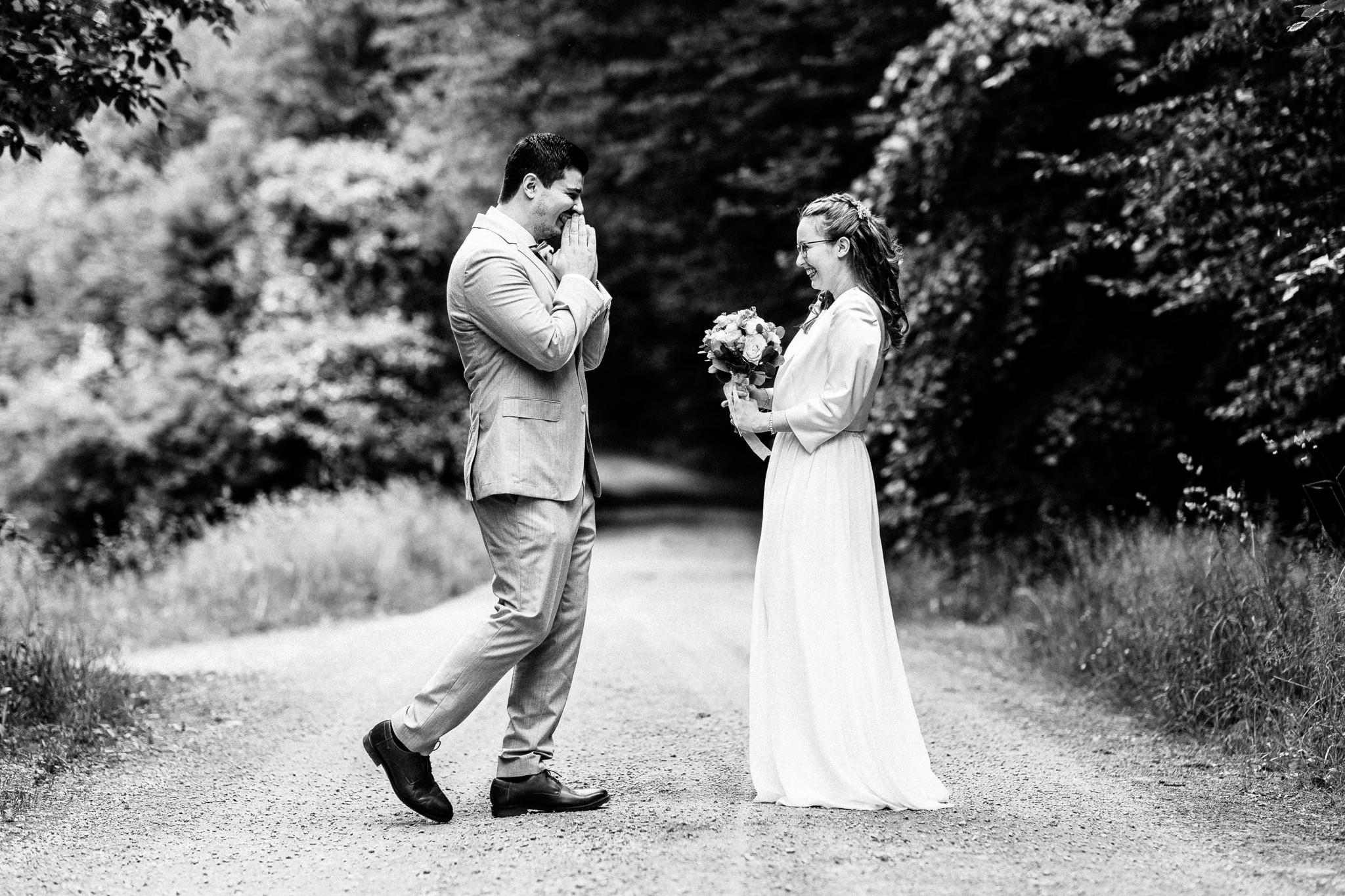 First Look Shooting vor der Trauung: In einem schwarz-weißen Bild steht der Bräutigam in hellem Anzug auf einem von Bäumen gesäumten Weg und hält sich aufgeregt die Hände vor’s Gesicht, während die Braut im schlichten, fließenden weißen Kleid lächelnd einen Blumenstrauß präsentiert.