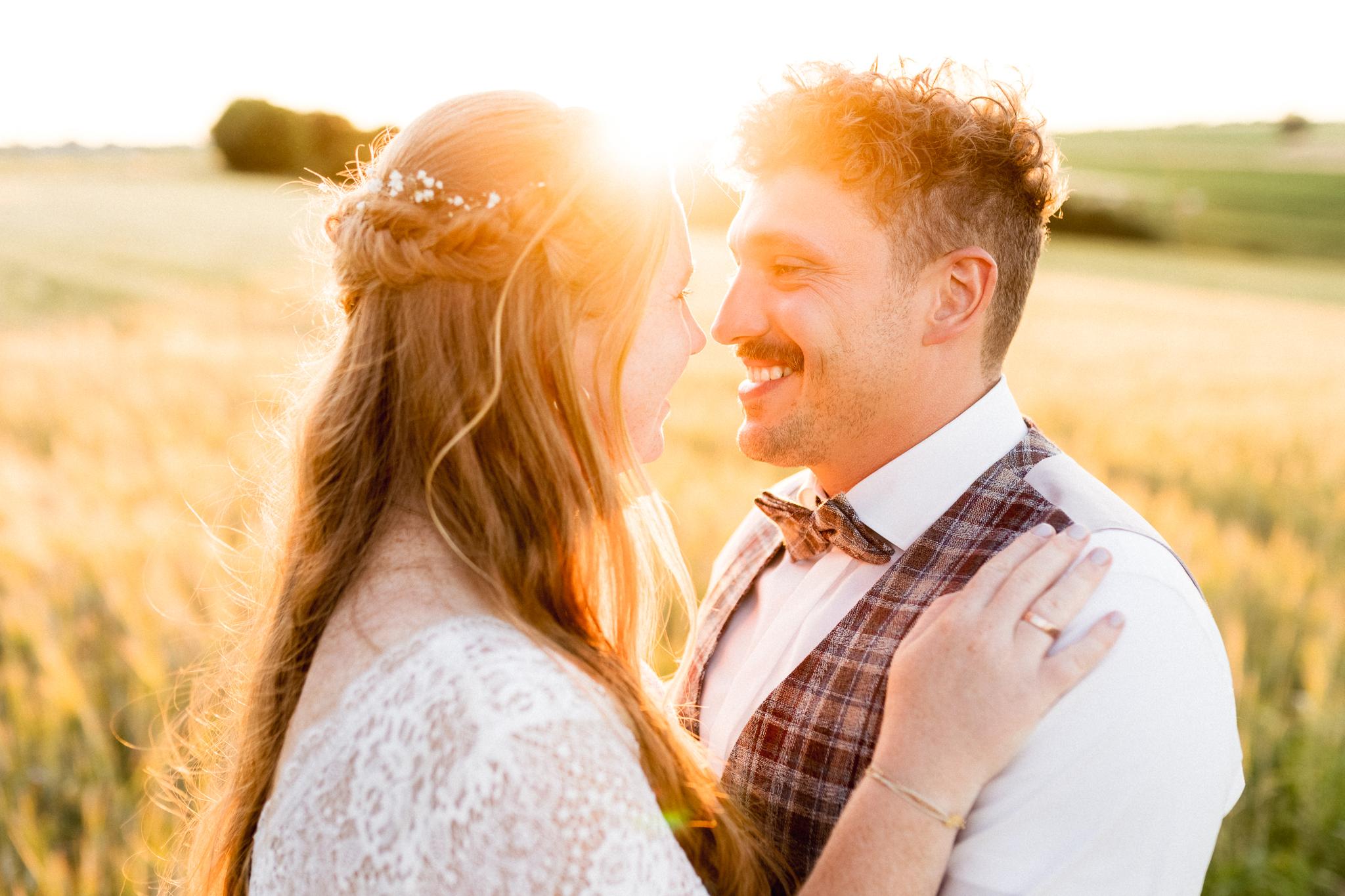 Brautpaarshooting beim Sonnenuntergang: Braut im Spitzenkleid mit locker geflochtenem Haar und Bräutigam in kariertem Gilet und Fliege stehen in einem goldenen Weizenfeld bei untergehender Sonne und lächeln sich verliebt an.