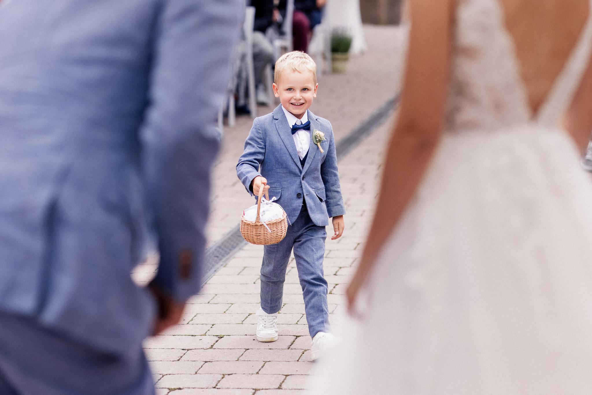 Kleiner Junge im eleganten Anzug bringt dem Brautpaar die Ringe während der freien Trauung in der Oase Weil, lächelnd und mit einem Körbchen in der Hand.