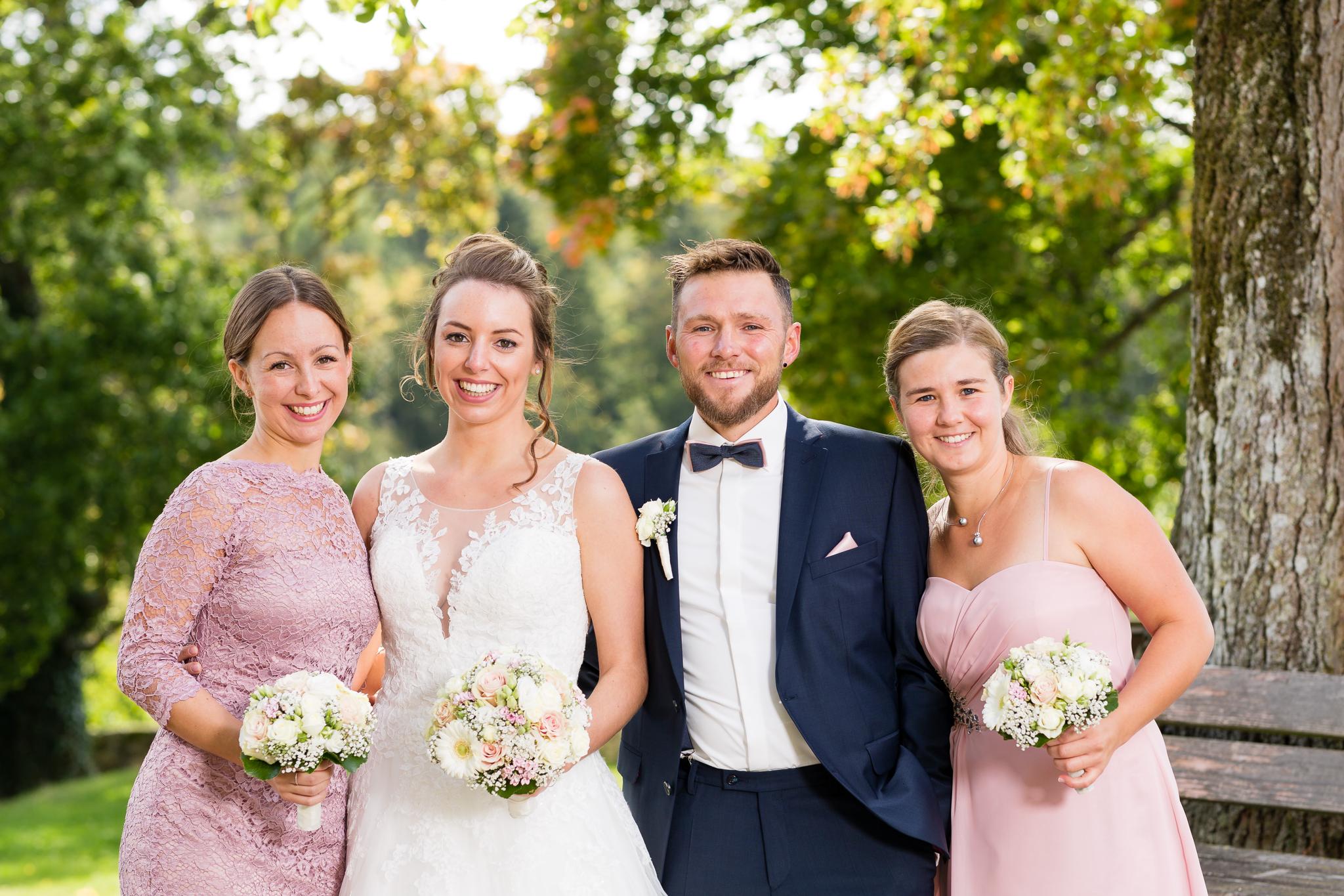 Braut und Bräutigam mit zwei Begleiterinnen im Garten: Die Braut im weißen Spitzenkleid, der Bräutigam im dunklen Anzug, beide flankiert von zwei in Pastelltönen gekleideten Freundinnen, alle lächeln und halten liebevoll ihre Brautsträuße.